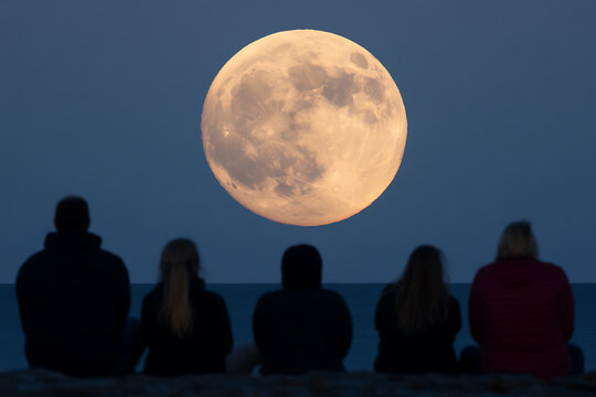 Silhouetted figures gaze at the moon. A group of friends admire a bright, round moon over the ocean. Serene moment on a beach, connecting with nature.
