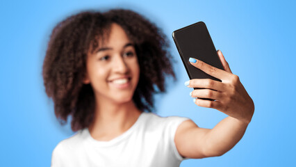 Fototapeta premium Photo for modern blog or social networks. Happy millennial african american lady in pink t-shirt with curly hair, makes selfie, focus on phone, isolated on violet background, studio shot, empty space