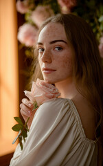 Obraz premium Light-skinned young woman, with freckles, holding a pink rose, posed indoors, romantic concept.