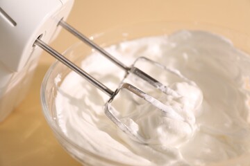 Hand mixer with whisks and bowl of whipped cream on beige background, closeup