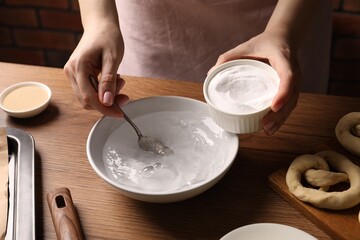 Making pretzels. Woman adding baking soda into bowl of water at wooden table, closeup