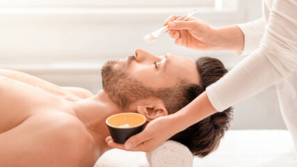 Handsome bearded man having face treatment at male spa salon, side view, copy space. Relaxed middle...