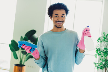 Smiling young man cleaning indoors with spray bottle and cloth showcasing a cheerful attitude and...