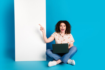 Young woman with curly hair sitting on blue background pointing excitedly at blank space
