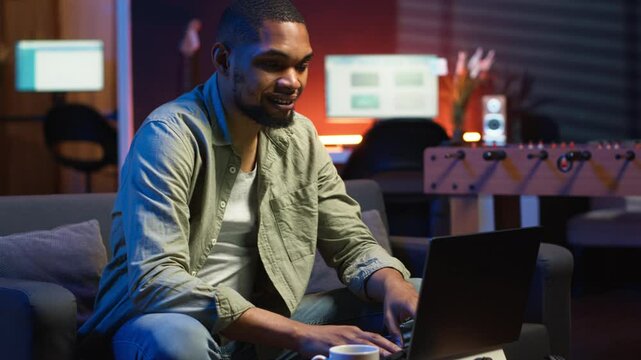 Young black adult working on his freelance tasks in the living room, adjusting his job schedule and enjoying work from his cozy home. Person navigating on web with laptop, multitasking. Camera B.
