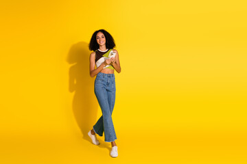 Stylish young woman holding a smartphone brightly smiling in front of vibrant yellow background...