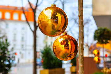 Two shiny gold Christmas ornaments hang from fir branch in urban setting, reflecting surrounding street and buildings. Blurred background features evergreen shrubs and a festive city atmosphere