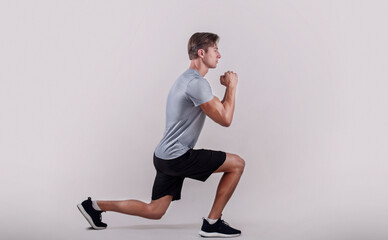 Side view of young fit guy in sportswear doing lunge on light studio background. Professional...