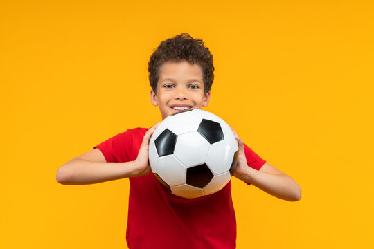 Studio shot of happy smiling adorable African American schoolboy wearing basic red t-shirt, posing with soccer ball in hands, isolated over bright colored orange yellow background