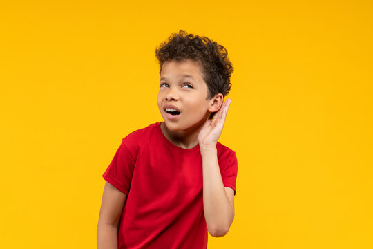 Adorable African American schoolboy posing over bright colored yellow studio background holding hand at his ear and listening to secret or gossip