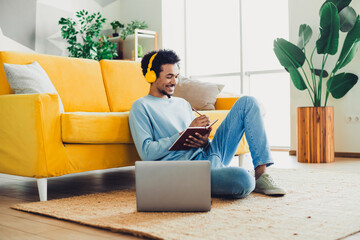 Young man wearing yellow headphones and blue sweatshirt sitting indoors with a notebook and laptop,...