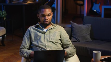 Portrait of african american adult typing data on his laptop at home, being self employed relaxing on a bean bag. Young person working remotely on freelancing tasks, casual teleworker. Camera B. - Powered by Adobe