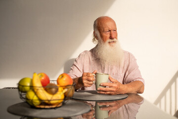 Elderly man with a long white beard sitting at a table with a cup of coffee and bowl of fresh fruits, enjoying sunlight on a peaceful morning