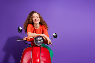 Smiling young woman on a red scooter, wearing a vibrant orange shirt against a purple background,...