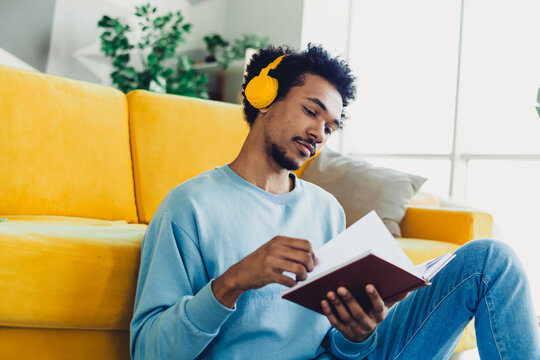 Young man relaxing at home wearing headphones and reading a book in a casual setting against a comfortable couch in a bright interior