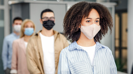 New normal and temperature check before work. Millennial multiracial employees in protective masks stand in line with social distancing at office in morning, free space, blurred, indoors