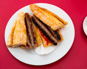 Baguette with sausage on white circular plate served on table with red tablecloth.