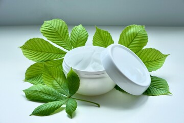 Jar of beauty facial or body cream with fresh green chestnut leaves on white table. Concept of natural herbal ingredients in cosmetic products.