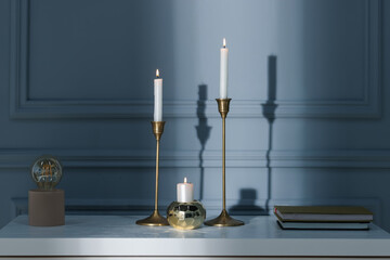 Stylish candlesticks with burning candles, books and lamp on white table near gray wall indoors