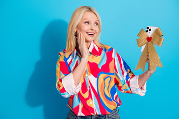 Smiling mature woman presents festive gift smartphone in colorful blouse against vibrant blue...
