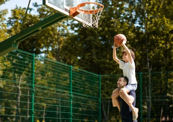 Fotobehang Happy Loving Family. Portrait of young curly boy sitting on father's shoulders, playing basketball, throwing ball to basket, man standing on court outdoors and helping his son, selective focus © Prostock-studio