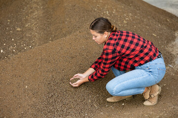 Young dairy farm female worker squatting at heap of soybean husk in fodder storage