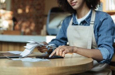 Small business management and accounting. Busy african american young lady owner in apron works with papers, counts costs and profits in interior of modern cafe, cropped, close up, copy space