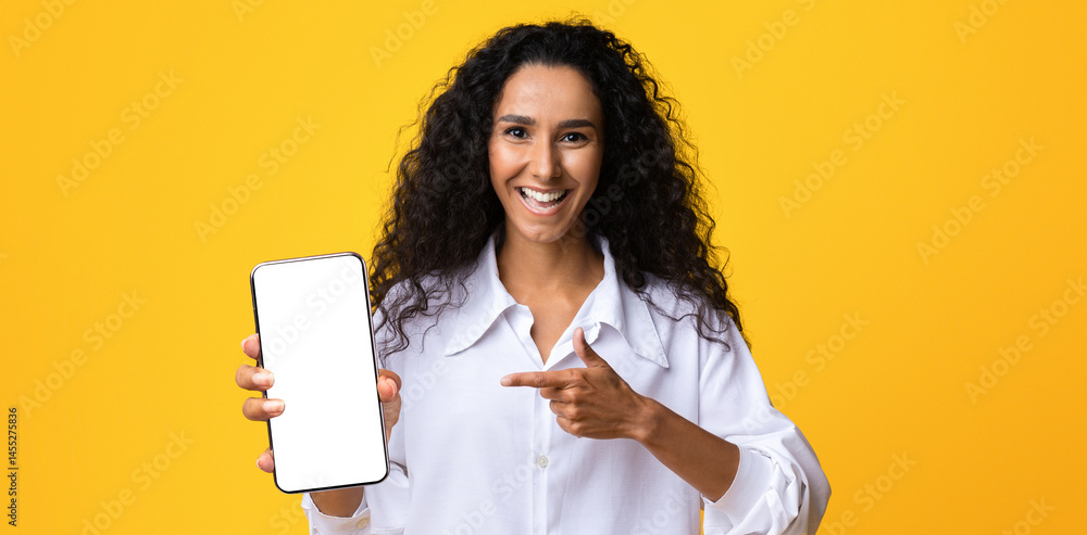 Poster joyful brunette lady pointing at smartphone with black blank screen, showing new app, isolated on ye