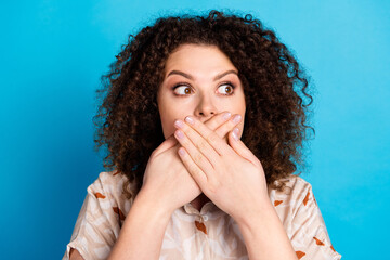 Young woman with curly hair covering her mouth with both hands, expressing surprise or shock,...
