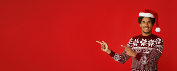 Recommendation for New Year and Christmas. Happy young african american man in santa claus hat and sweater with pattern points fingers at free space, isolated on red background, panorama, cut out