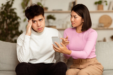 Jealous Korean Wife Showing Cheating Husband His Smartphone Demanding Explanation Sitting On Couch At Home. Marital Problems, Jealousy And Infidelity In Relationship Concept