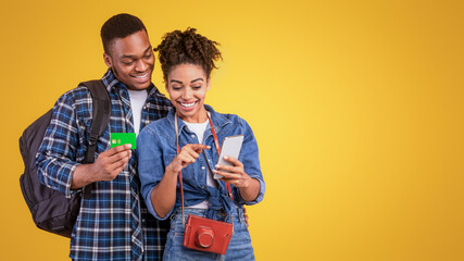 Happy Black Couple Booking Travel Tickets Online Using Smartphone And Credit Card Standing With Suitcase Over Yellow Studio Background. Mobile Banking Abroad, Traveling Budget And Finances.