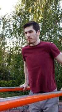 A young man in a T-shirt and pants performing an exercise on parallel bars outdoors. A strong, focused workout routine showing body strength and fitness training
