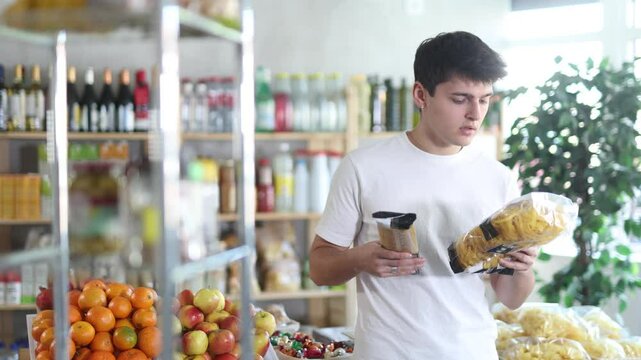 Thoughtful young guy holding two different pasta packages and pondering over meal choice for dinner during shopping in grocery store, evaluating ingredients and nutritional value before purchasing 
