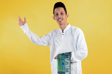 Smiling muslim man wear white shirts with Islamic ornaments while dangling a prayer mat in his hand...