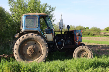 Obraz premium An old blue tractor stands in a green meadow near trees, its paint partially peeled off, revealing rust, with a cultivated field visible in the background