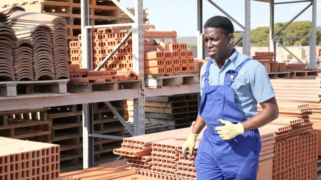 Concentrated afro american man worker stacking bricks on pallet in warehouse of building materials
