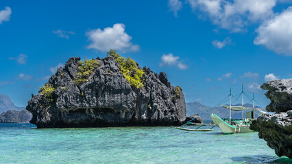 The traditional Filipino bangka boat is anchored in the aquamarine ocean next to a small rocky...
