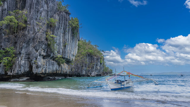 A traditional Filipino bangka boat decorated with colorful flags is moored near the shore. The ocean waves are foaming on the sand. Green tropical vegetation on the steep slopes of the coastal cliff. 