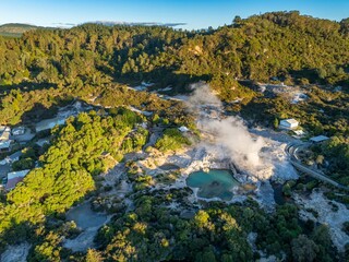 Aerial view of the Wai-O-Tapu geothermal area in New Zealand. Steam rises from the hot springs, surrounded by lush vegetation and a boardwalk for visitors. A popular tourist destination.