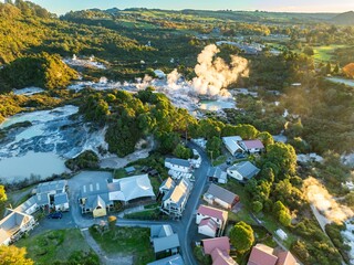 Aerial view of Whakarewarewa Thermal Village, Rotorua, New Zealand. Steam rises from geothermal pools, a natural wonder attracting tourists. Houses are nestled near the park.
