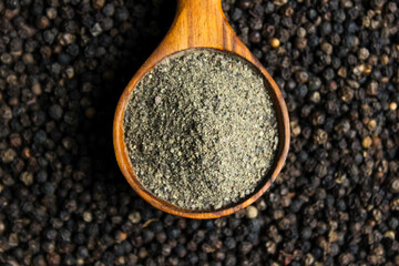 Black pepper powder in wooden cup spoon with black peppercorns background. Food ingredients. Aromatic spice. Top view