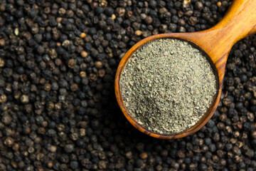 Black pepper powder in wooden cup spoon with black peppercorns background. Food ingredients. Aromatic spice. Top view