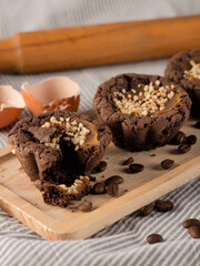 Brown muffins on wooden tray among coffee beans, eggshell, rolling pin on striped fabric, close-up, vertical image.
