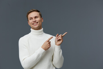 Bright smiling young man with short brown hair wearing a cozy white turtleneck sweater pointing to...