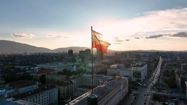 A close-up view of the flag of Bulgaria blowing in the wind against a sunset or sunrise over the capital city of Sofia. The Bulgarian flag flying over the Parliament building.