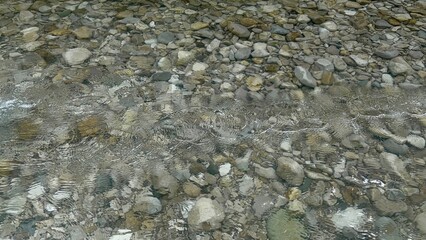 Clear water flows gently over smooth stones in a serene riverbed. Sunlight glimmers on the surface, creating a peaceful atmosphere in nature
