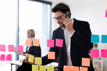 Young adult male using colorful sticky notes for business task organization on glass board. Holding phone engaged in conversation in modern office setting.