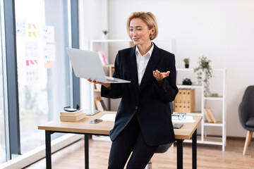 Confident middle-aged caucasian female dressed in business attire conducting video call in office...