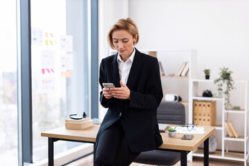 Professional businesswoman in modern office leaning on desktop browsing phone, wearing formal...